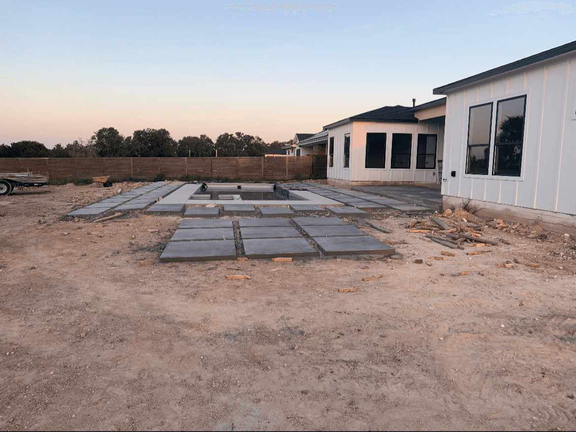 Square concrete pavers being installed around a swimming pool behind a white modern farmhouse.